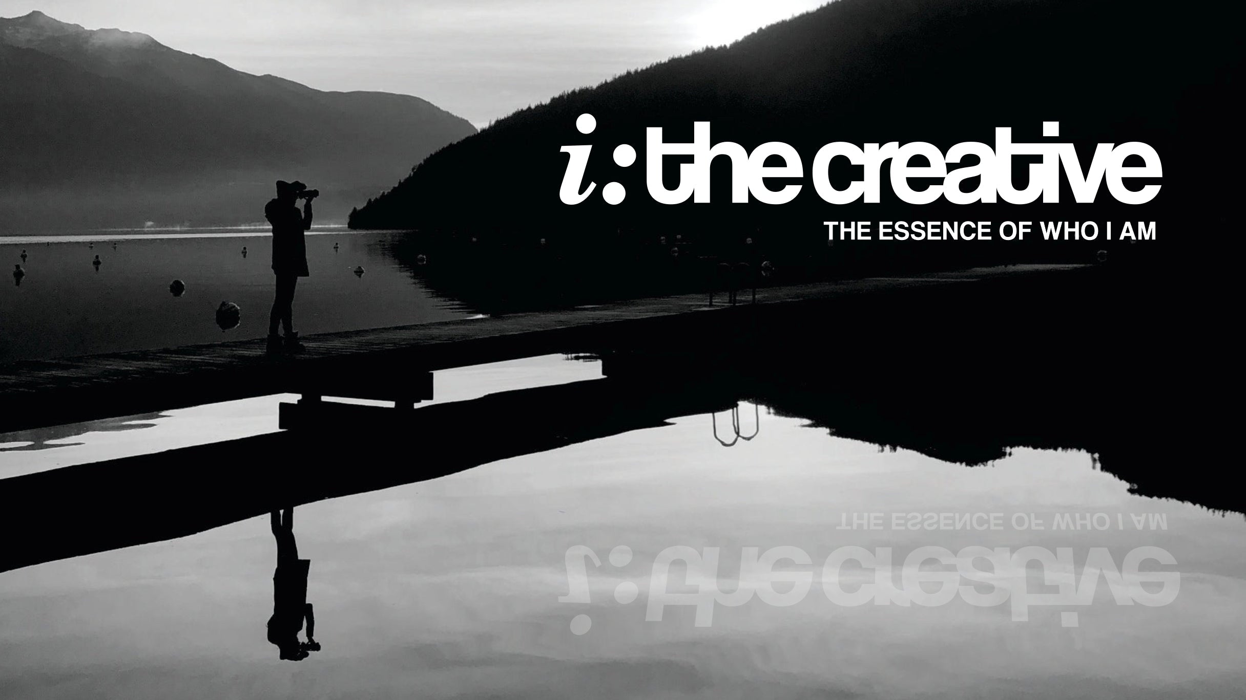 The silhouette of a photographer stands on the dock taking a photo of the black shadow of a mountain. The reflection mirrored on the silver lake. The white words i: the creative, THE ESSENCE OF WHO I AM superimposed on the mountainside faintly reflect on the water.