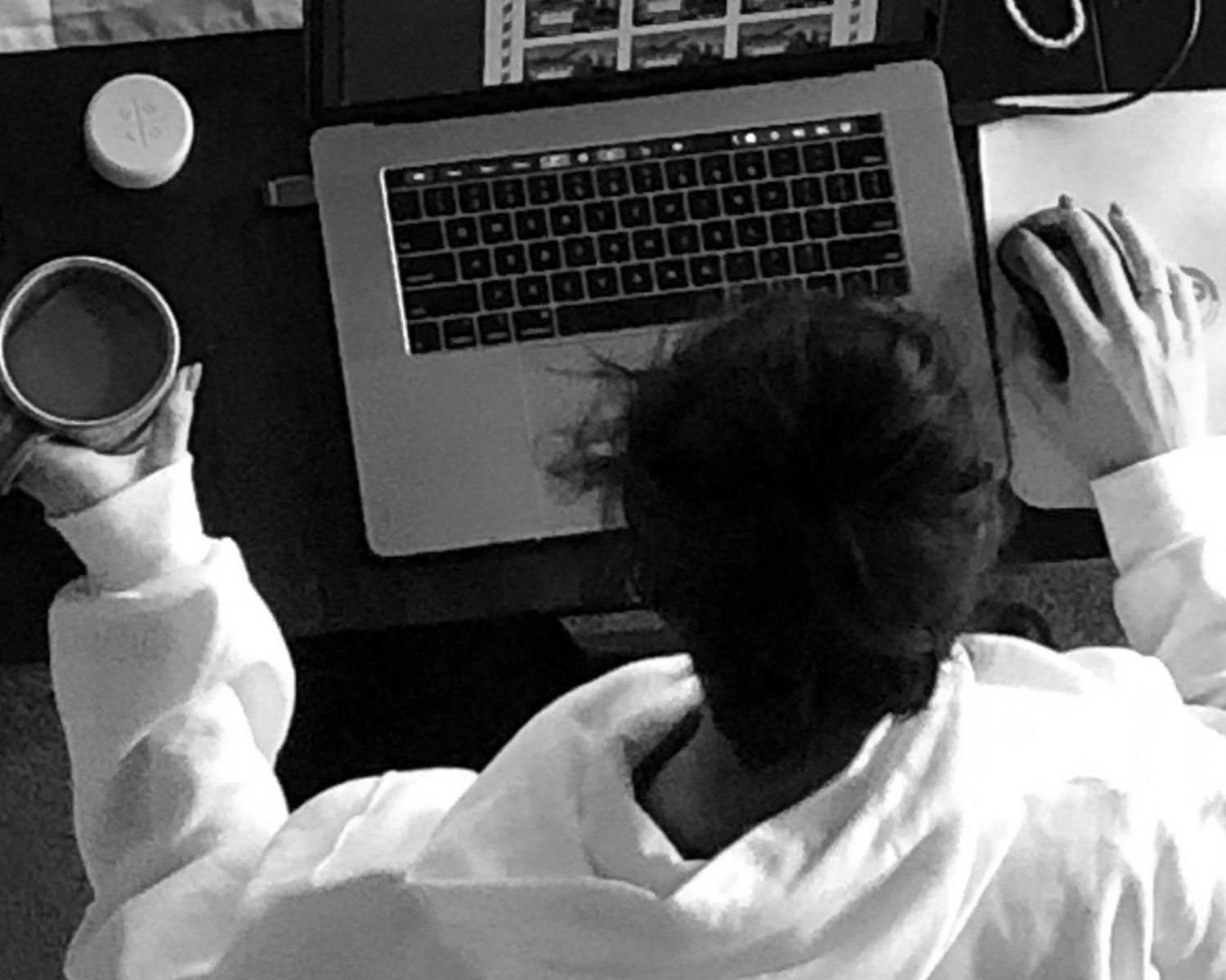 This black and white overhead view of a girl, hair up in a bun, wearing a white hooded sweatshirt, sitting at her silver computer, hand on the mouse, other hand holding her coffee cup.