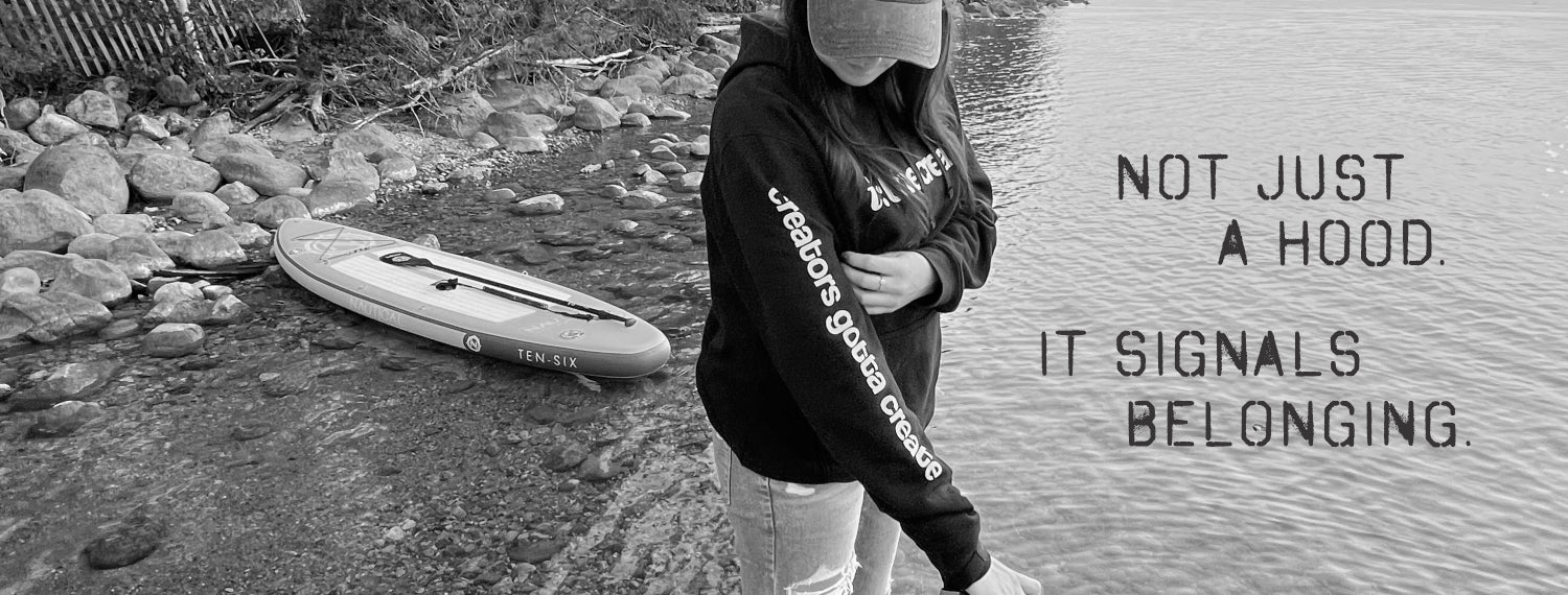 A black and white lifestyle photograph of a person wearing a black hoodie with 'Urbano Global Designs' printed on the sleeve, standing by the water next to a paddleboard. The text on the right reads 'NOT JUST A HOOD.
