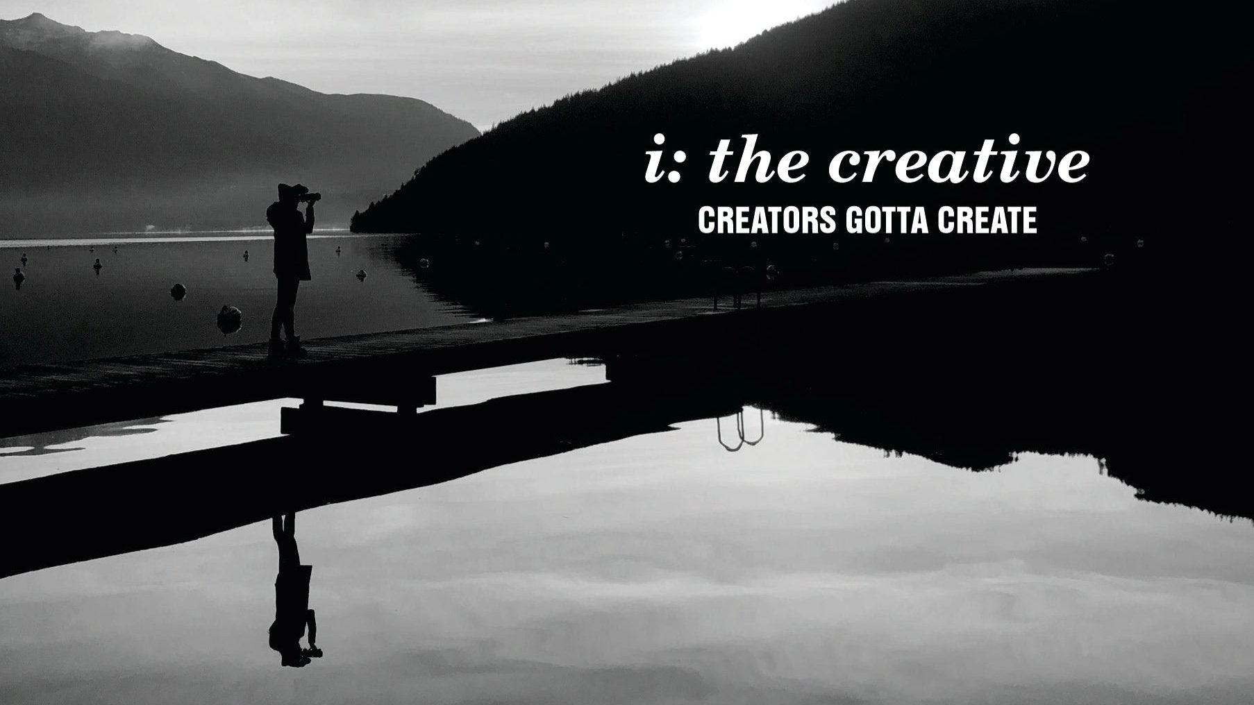 This is a black and white image depicting a silhouette of a photographer standing on a grey silhouetted dock, aiming at the black silhouette of a mountain against a cloudy grey sk. The whole scene is reflected on the mirror like silver lake. The brand name i: the creative and mott CREATORS GOTTA CREATE is superimposed against the black mountain.