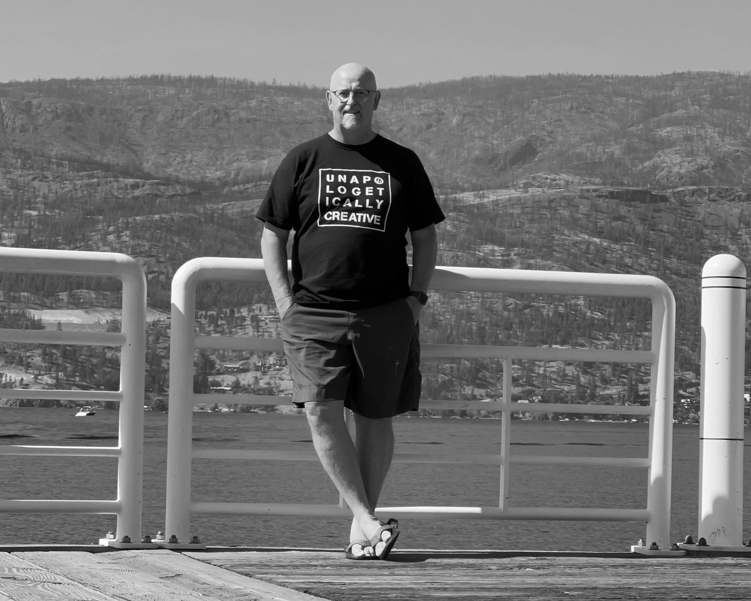 The bald owner of the brand stands to the right, wearing a black t-shirt with a white square design with word UNAPOLOGETICALLY broken into 3 lines and then CREATIVE as the fourth line. Behind him is a large white pipe railing against Okanagan Lake. The grey mountainside is in the distance and is sparsely covered with pine trees, half of which are burnt. The white bold quote is on the upper left of the image. 