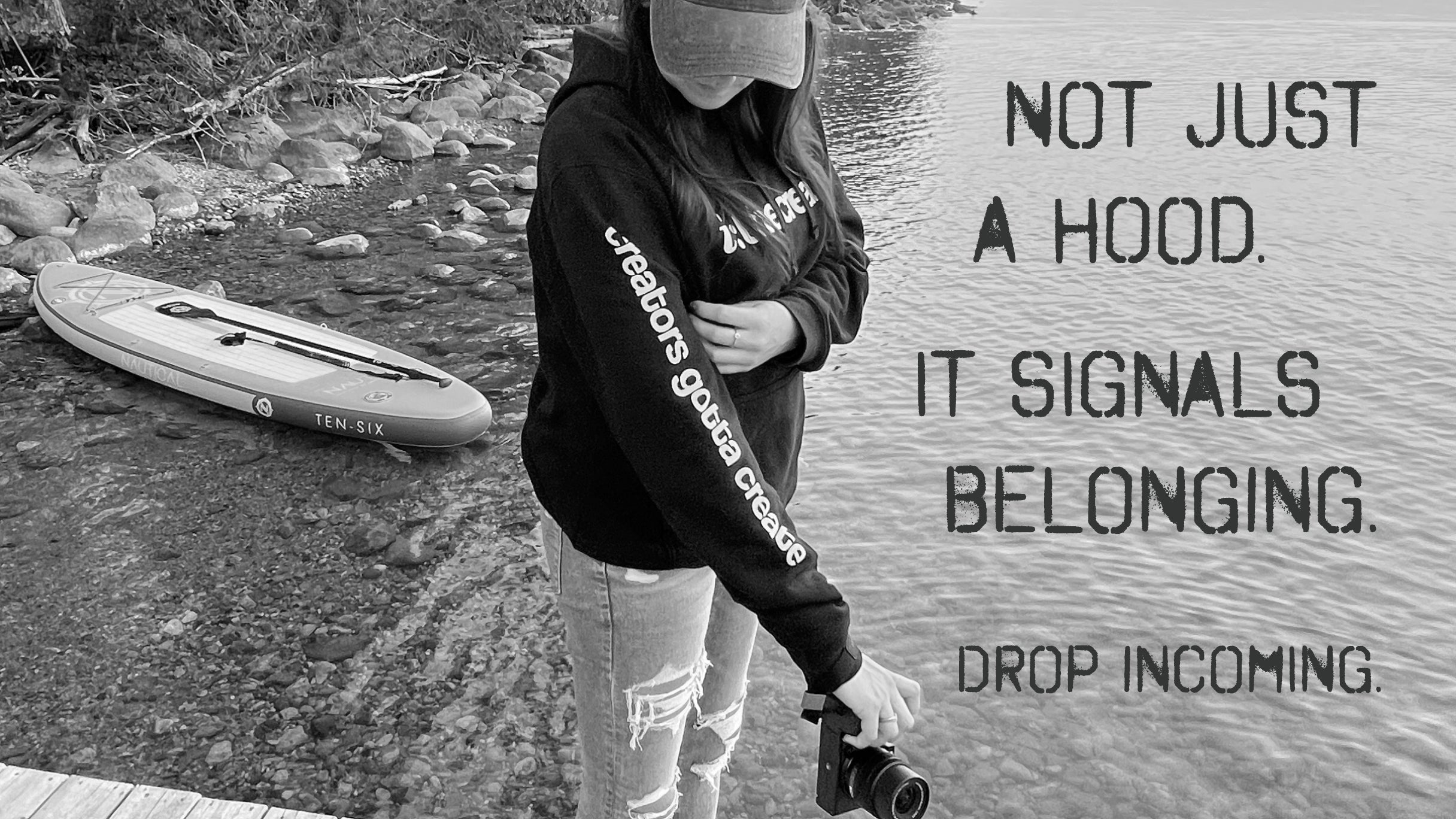 Person with a black hoodie with white lettering "creators gotta create" on the right sleeve. She holds a camera and a paddleboard sits on the rocky shoreline.